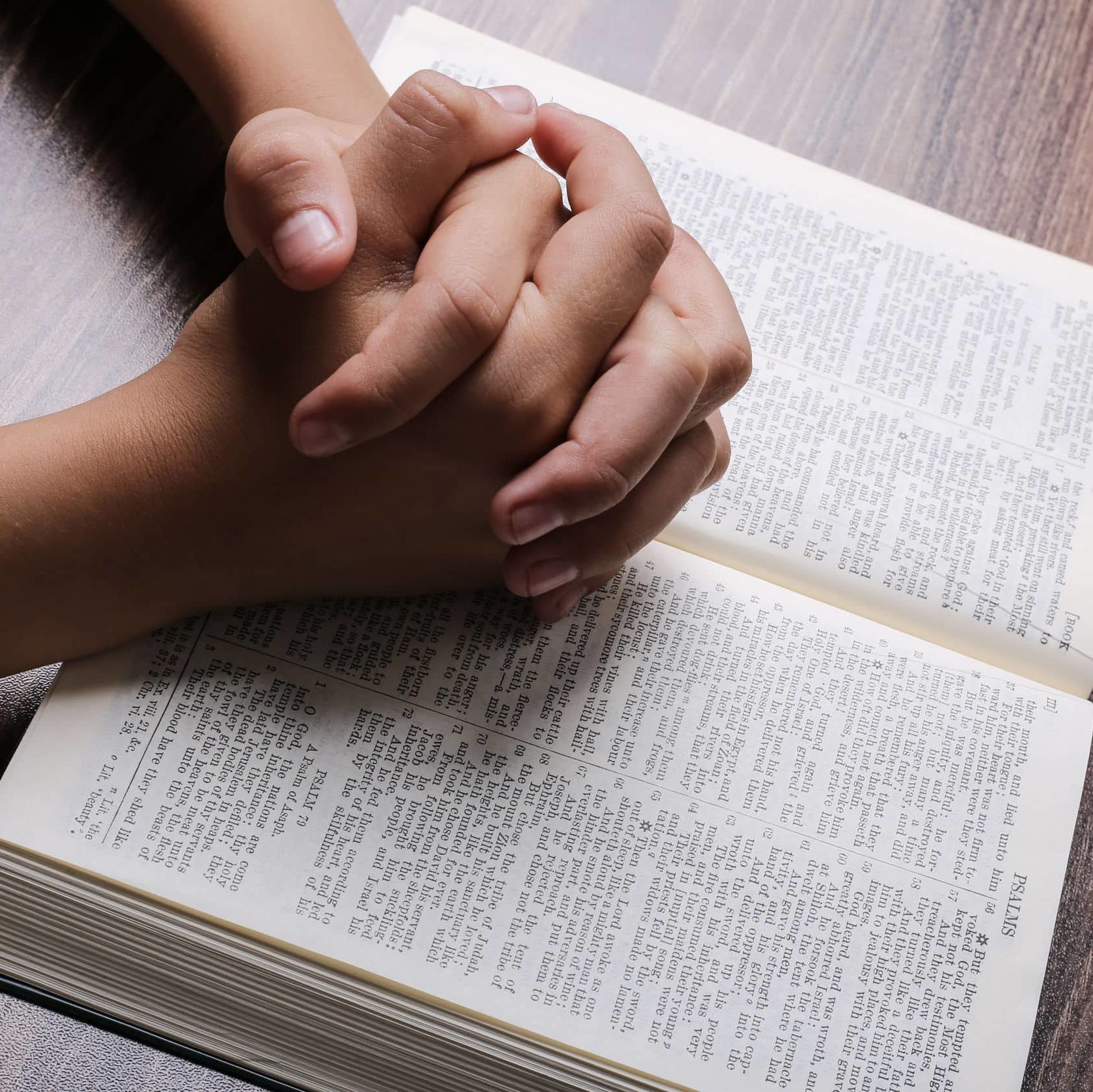 Praying hands with opened holy bible on the wooden desk.
